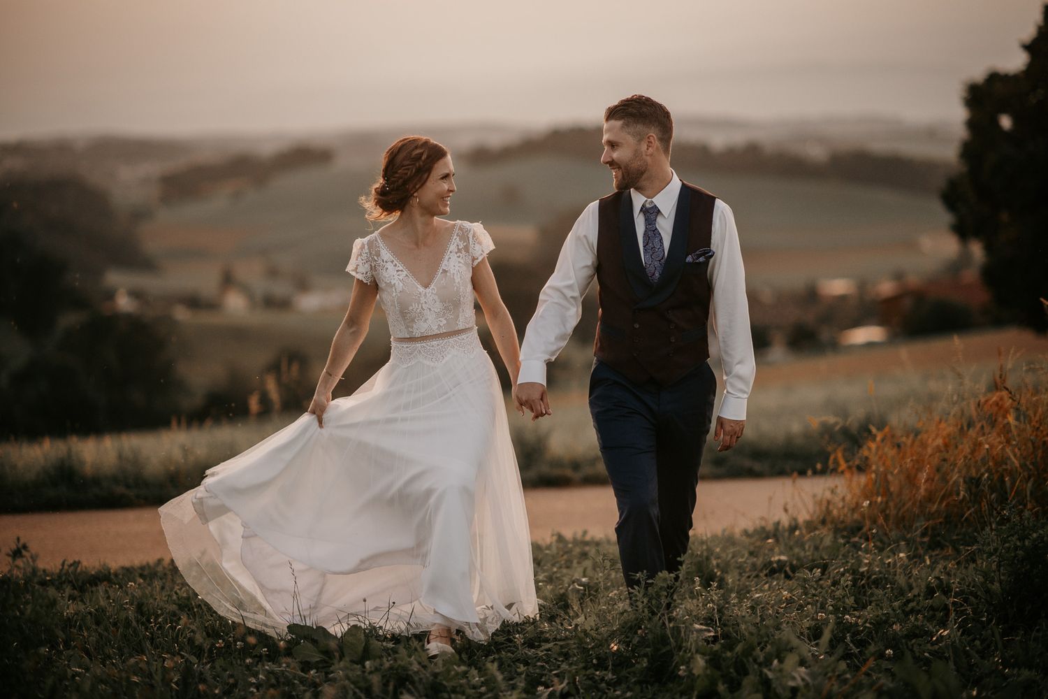 Bride and groom walking hand in hand through a scenic countryside at golden sunset, brides dress flowing.