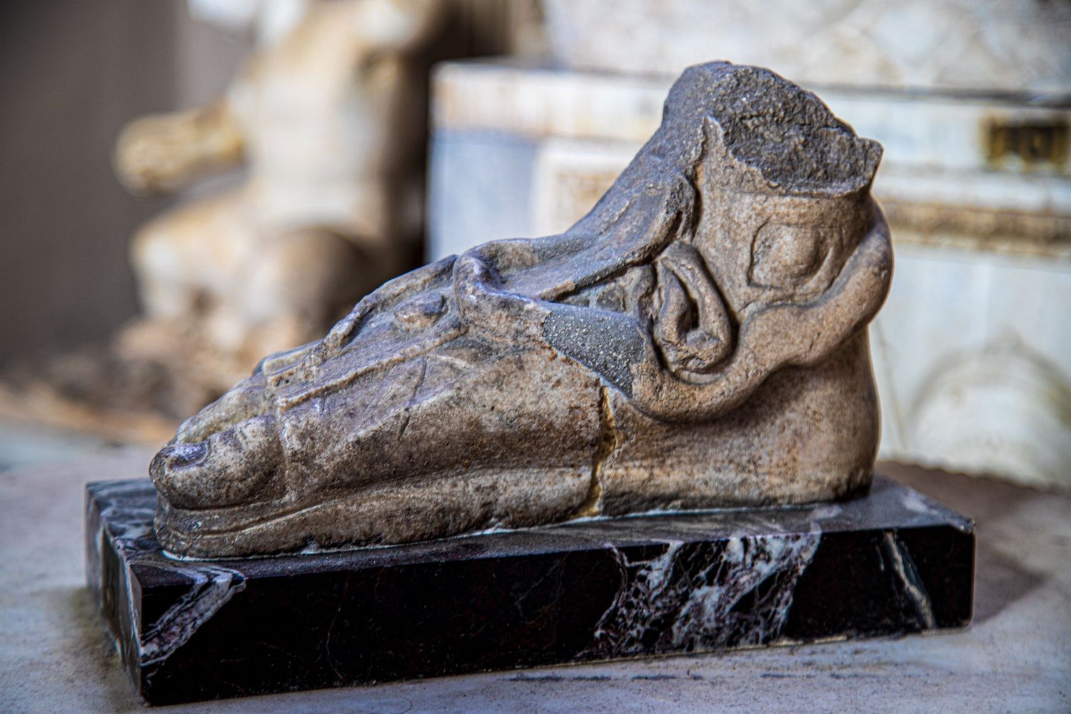 Ancient carved stone sculpture of a decorative foot or boot mounted on a black marble base.