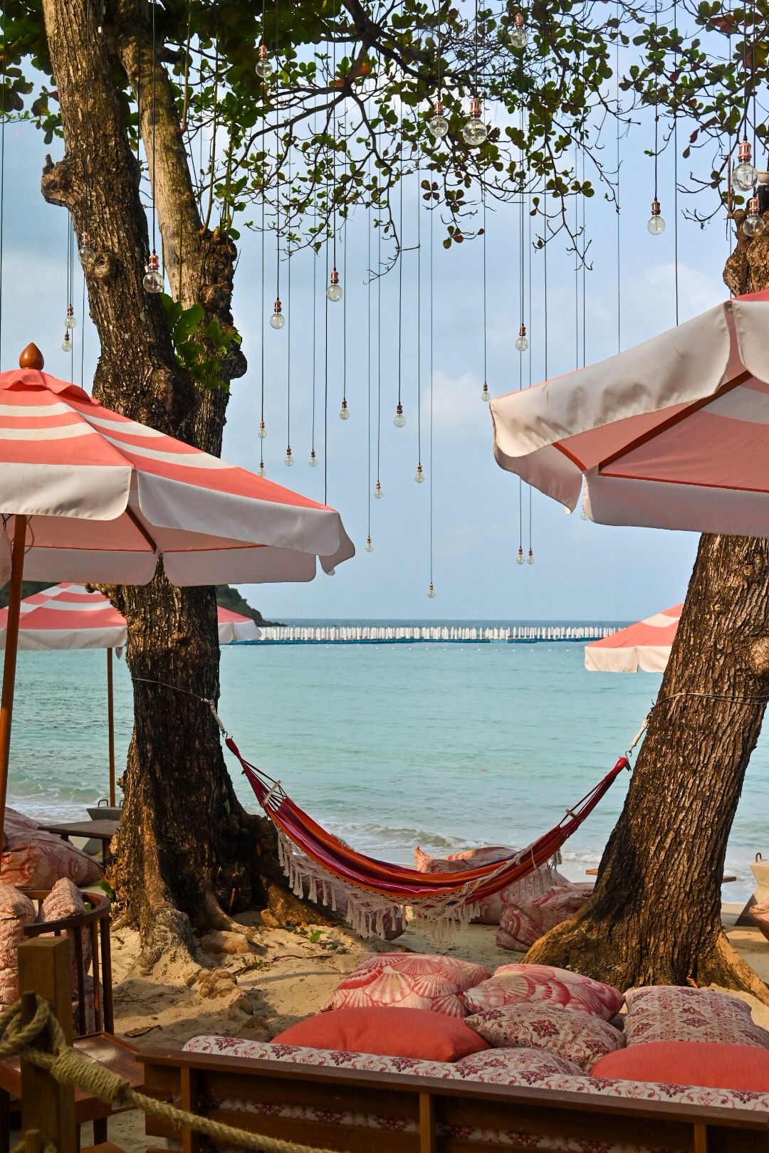 Beach hammock setup with coral umbrellas and string lights hanging from trees by turquoise waters.