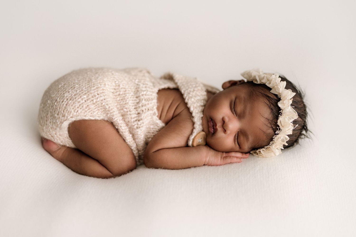 Sleeping newborn baby wearing white knit romper and floral crown lying on soft light backdrop.