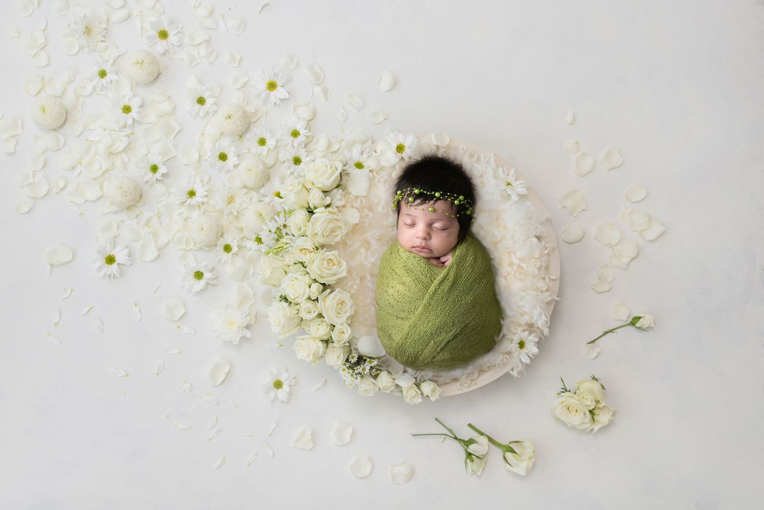 Overhead newborn photo with delicate white flowers scattered around a soft green wrap on a light background.