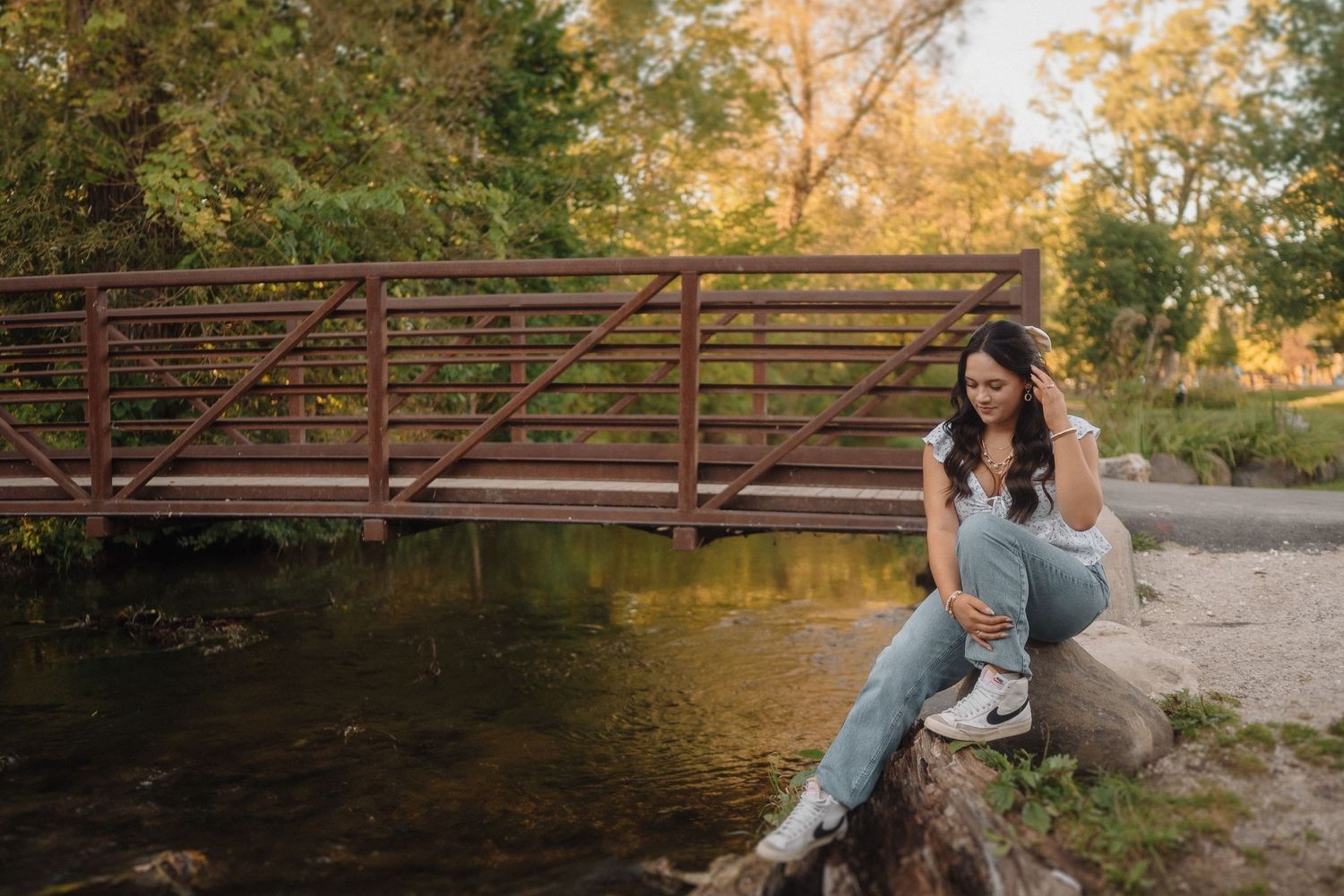 Someone in casual attire sits on the edge of a creek near a rustic metal bridge during golden hour sunset.