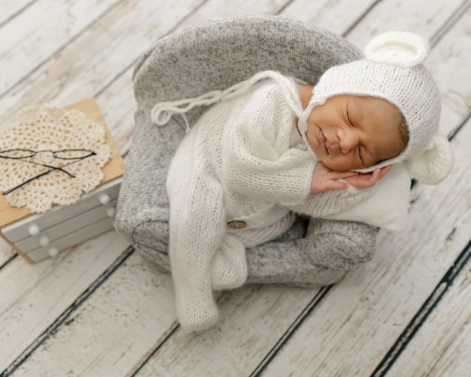 A sleeping newborn in white knit clothing on a wooden floor with neutral toned accessories.