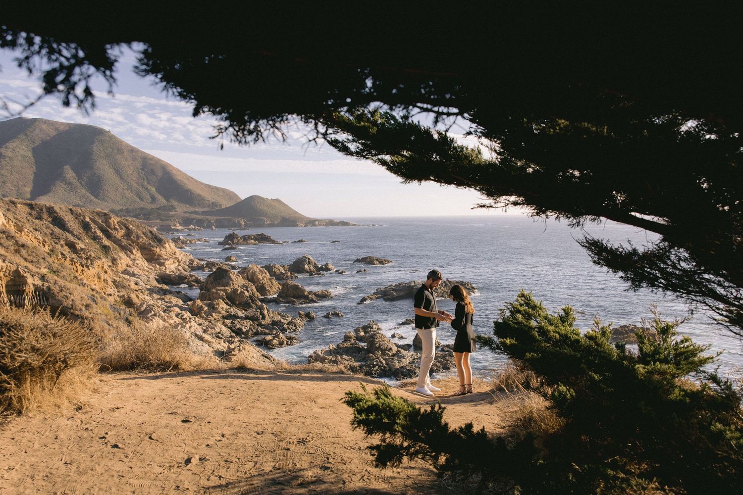 Scenic coastal viewpoint at Big Sur with figures overlooking rocky shoreline and mountains through cypress tree frame.