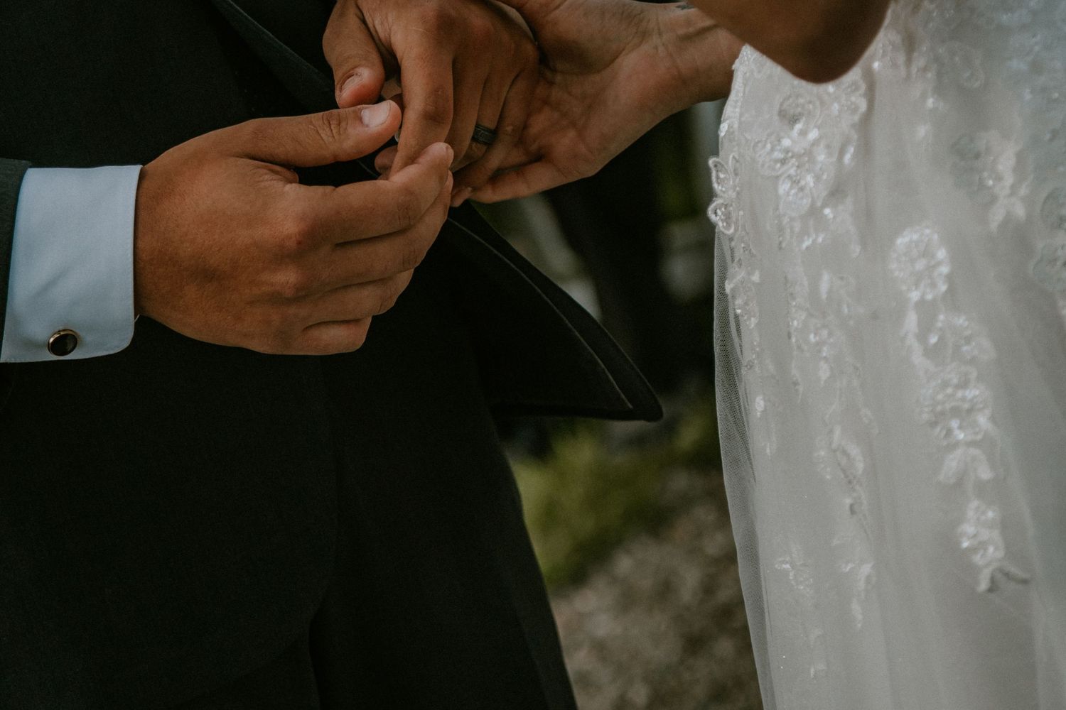 Close-up detail of hands holding wedding rings during outdoor marriage ceremony, with black suit and white dress visible.