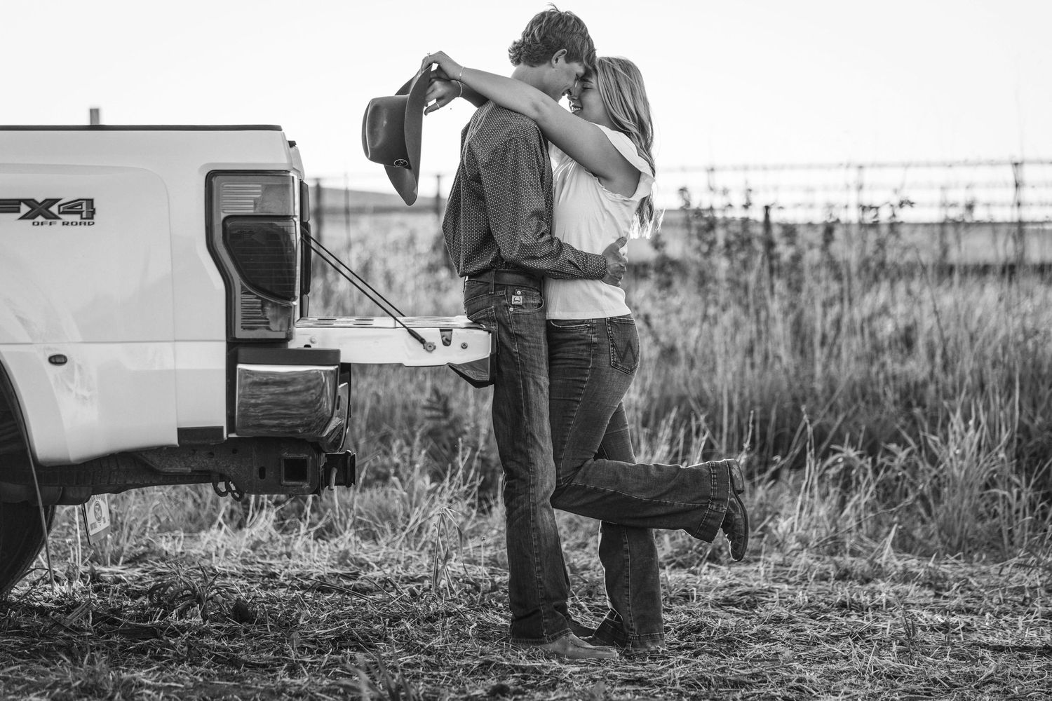 A black and white photo showing a romantic embrace next to a pickup truck in a grassy field.
