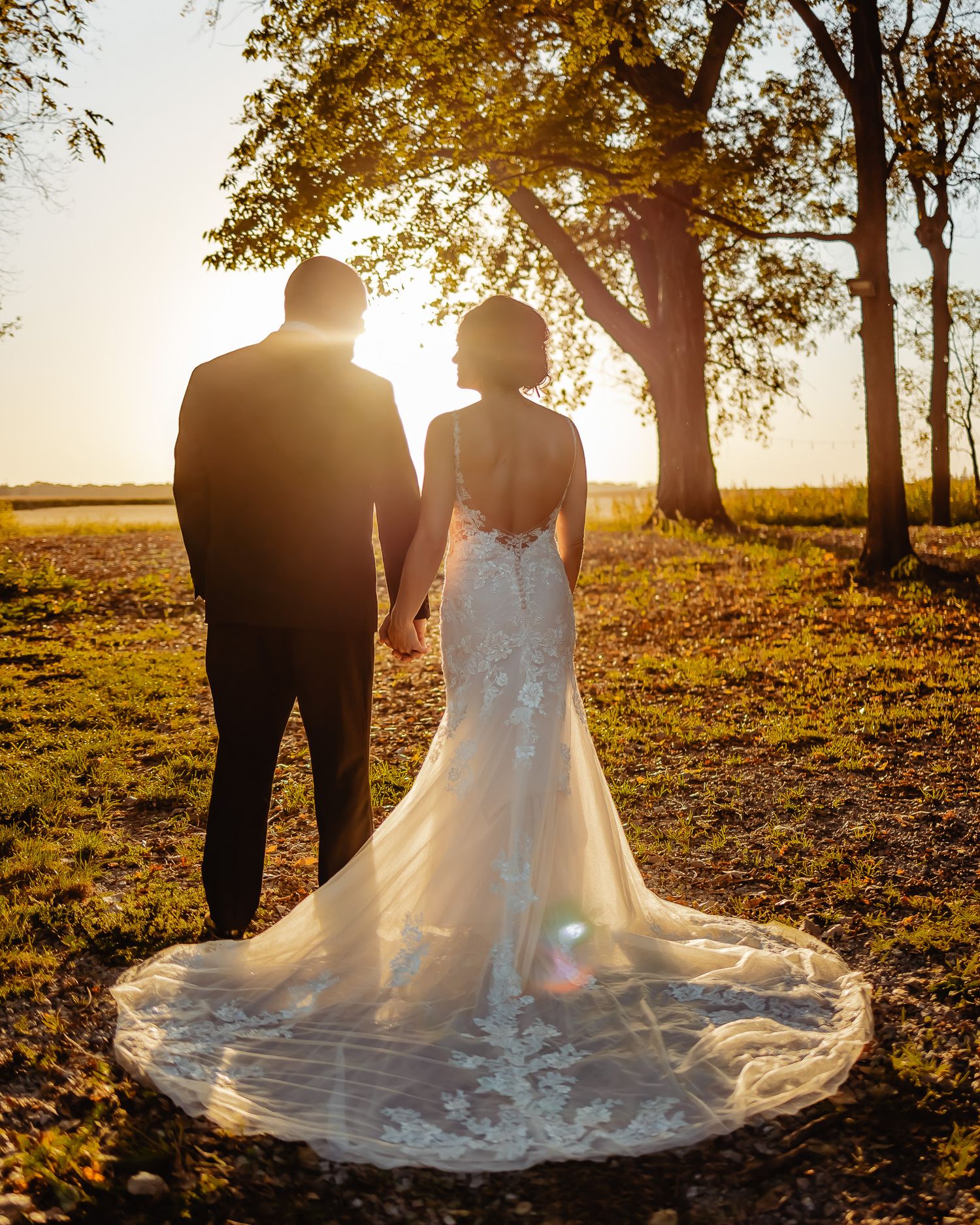 A couple stands in silhouette against a golden sunset beneath autumn trees while wearing elegant wedding attire.