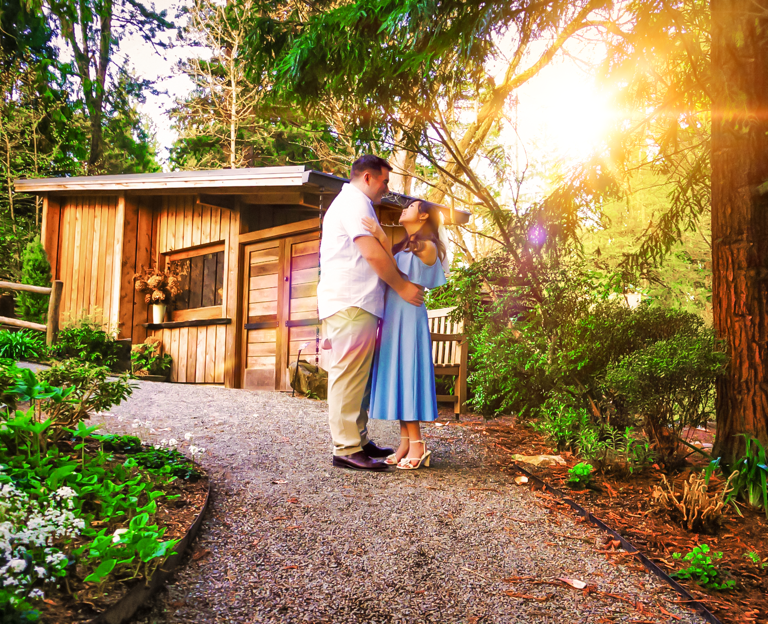 Two people embrace on a gravel path by a rustic cabin during golden sunset hour in a flower-filled garden.