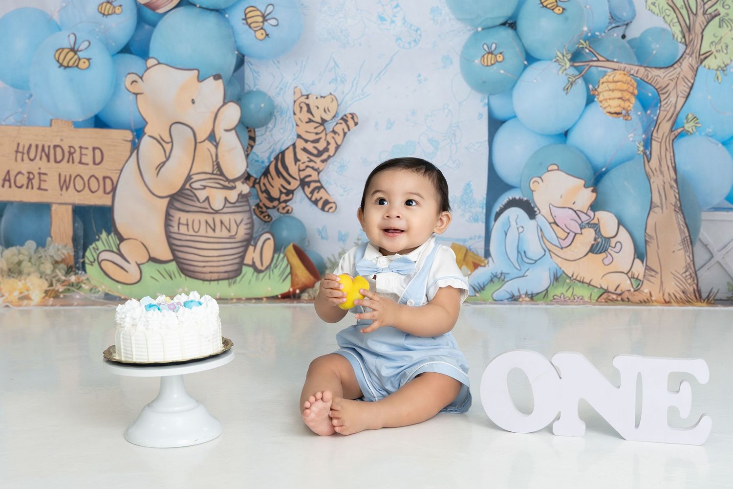 First birthday celebration setup with Winnie the Pooh themed backdrop, white cake on stand, and baby sitting with balloons.