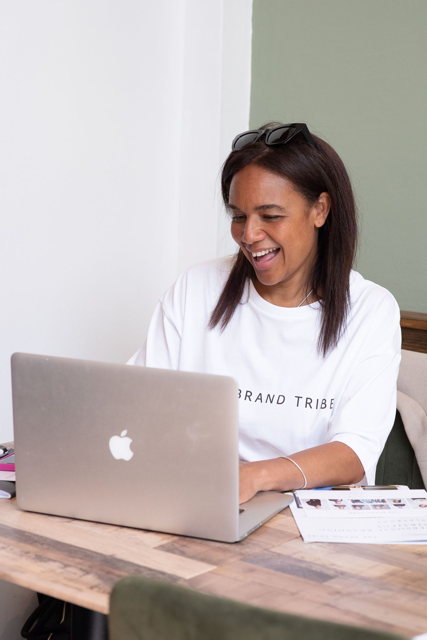 A person in a white t-shirt smiles while working on their MacBook laptop at a wooden desk.