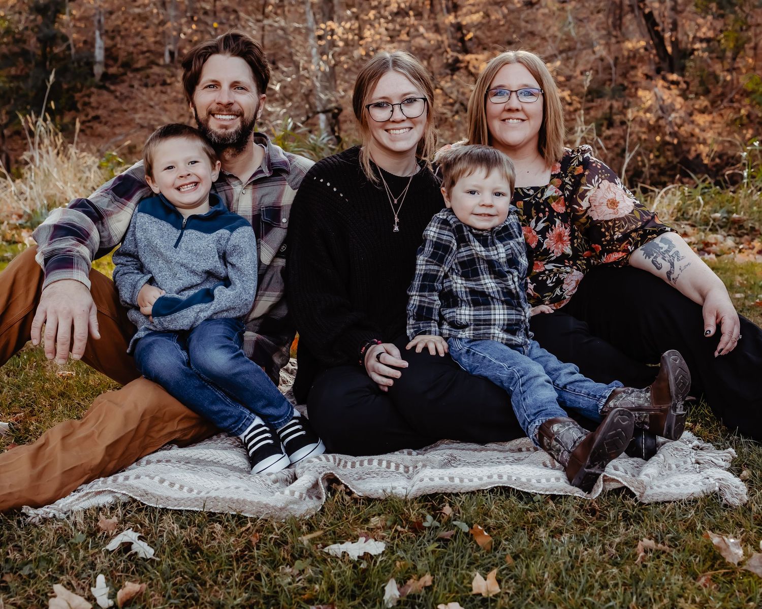 A family in casual plaid clothing sits together on a blanket outdoors during autumn with fallen leaves in the background.