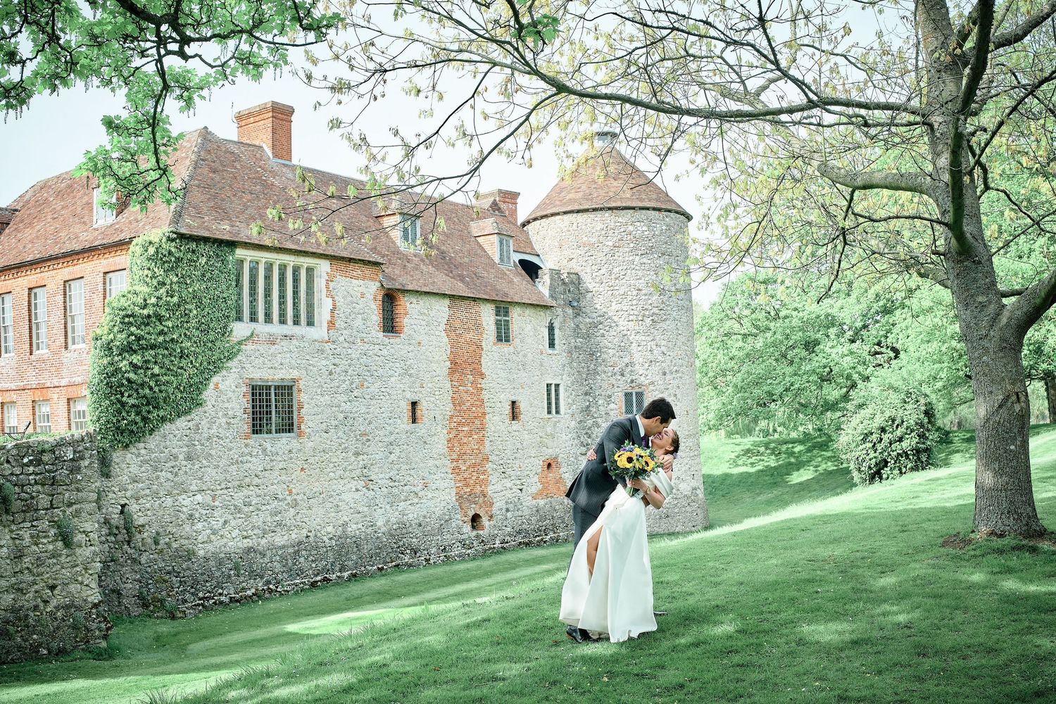 Elegant couple embraces on green lawn beside historic stone castle with ivy-covered walls and round tower during romantic moment.