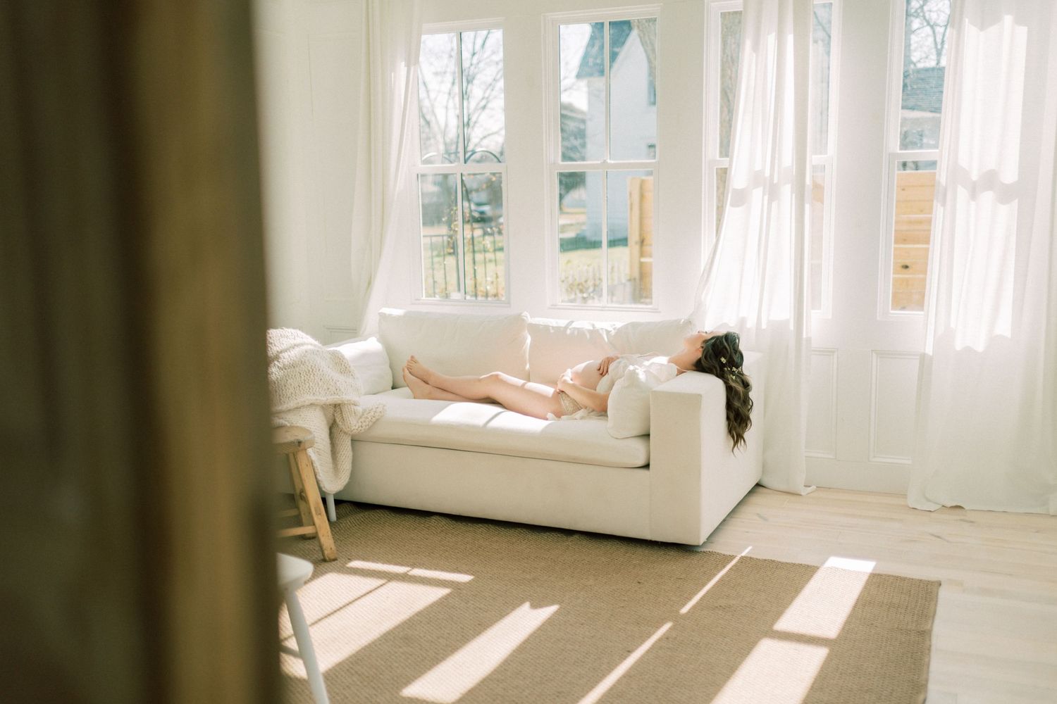 Sunlight streams through white curtained windows onto a cream couch in a bright, minimalist living room.