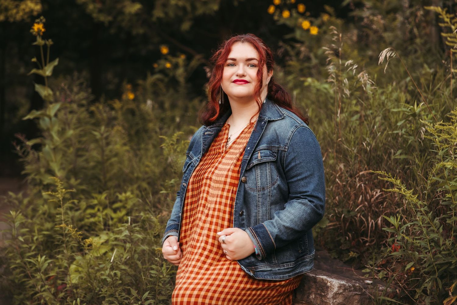 A high school senior wearing an orange plaid dress and denim jacket stands outdoors among tall grass and autumn foliage in Ann Arbor.