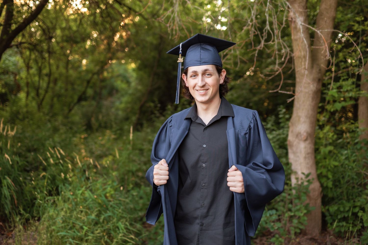 Graduate in navy blue cap and gown poses outdoors against a green forest background in natural lighting in Novi Michigan