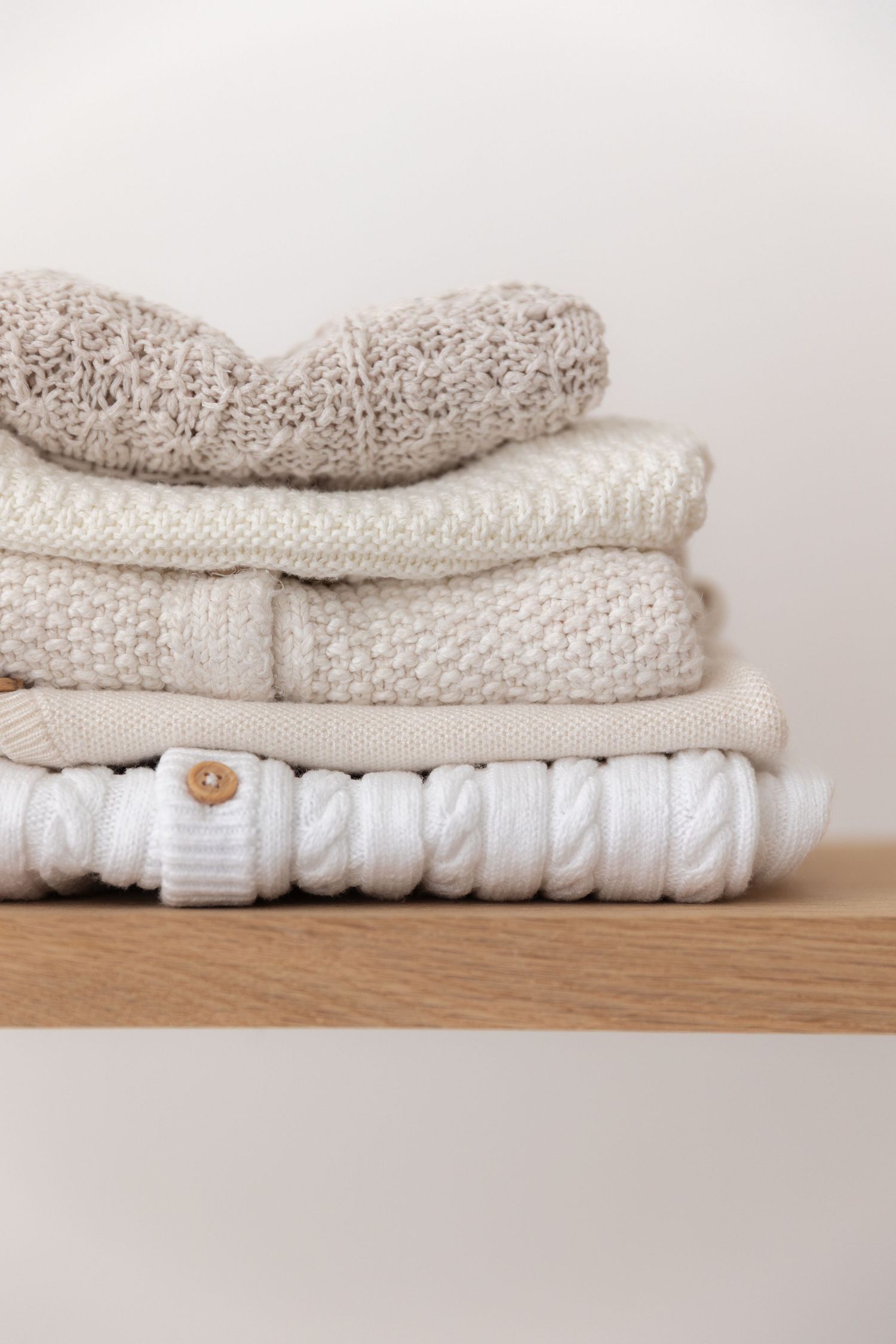 Stack of cozy knit sweaters in neutral cream and white tones with cable knit patterns on wooden shelf.
