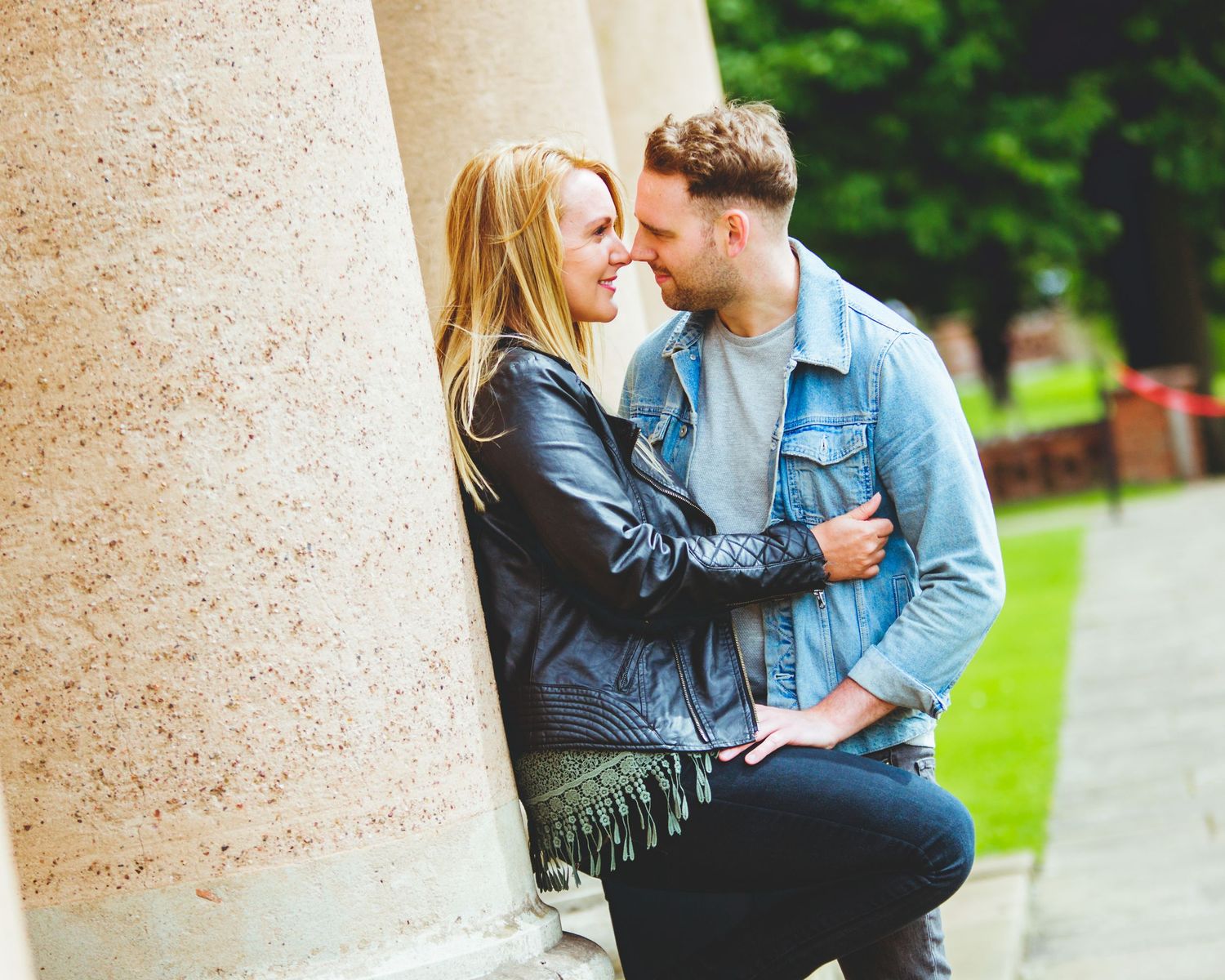 A romantic couple in casual jackets embraces against a stone pillar in a garden setting.