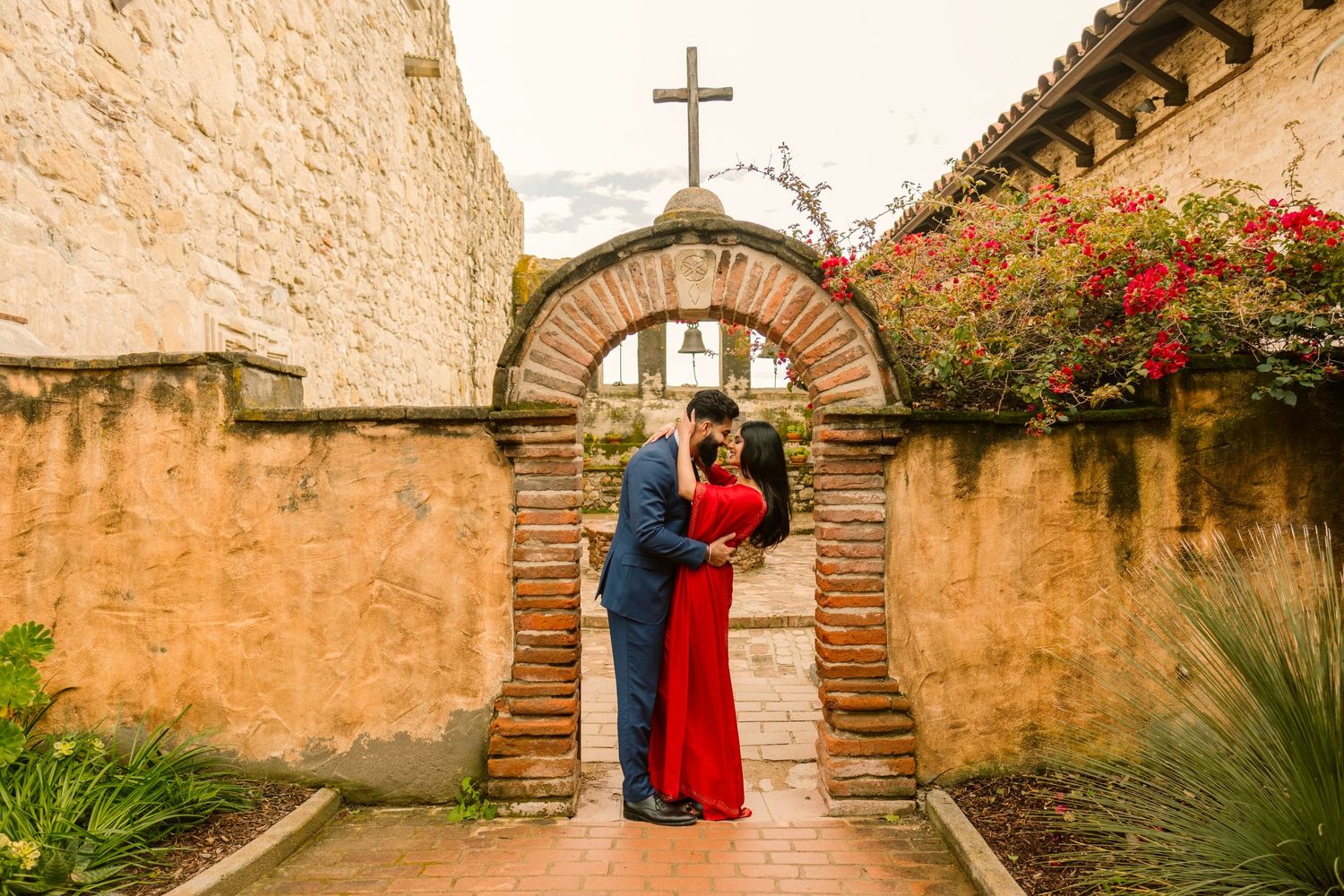 Couple embraces in romantic moment under stone archway with cross at Spanish mission adorned with red flowers.