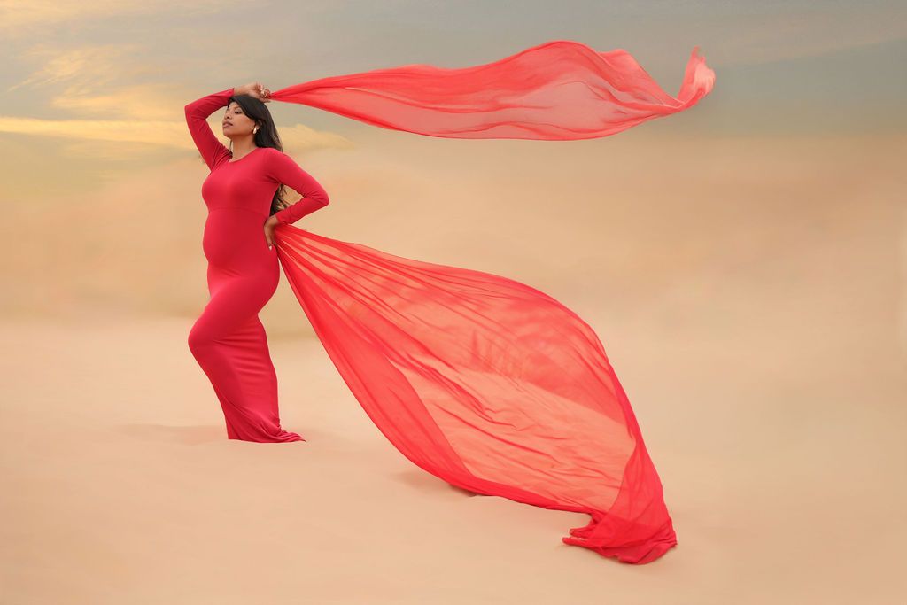 Dramatic maternity photo with flowing red fabric against sunset sky.