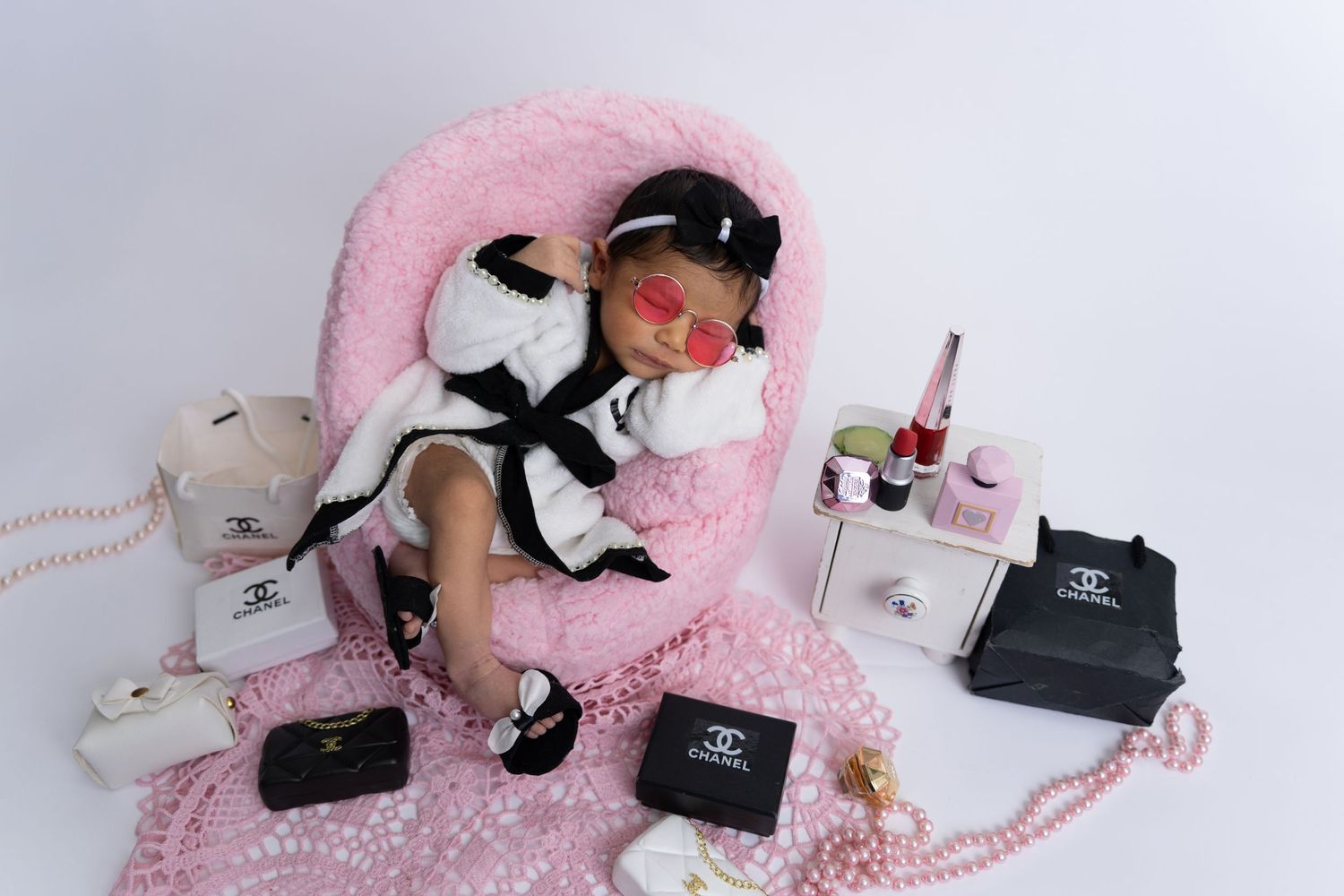 A person resting on pink carpet surrounded by luxury fashion items including handbags, perfume bottles, and accessories.