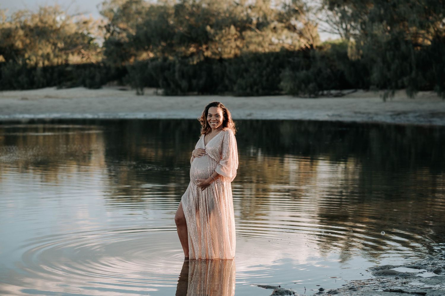 Elegant maternity photo at sunset shows an expectant mother in a flowing dress standing in calm lake waters.