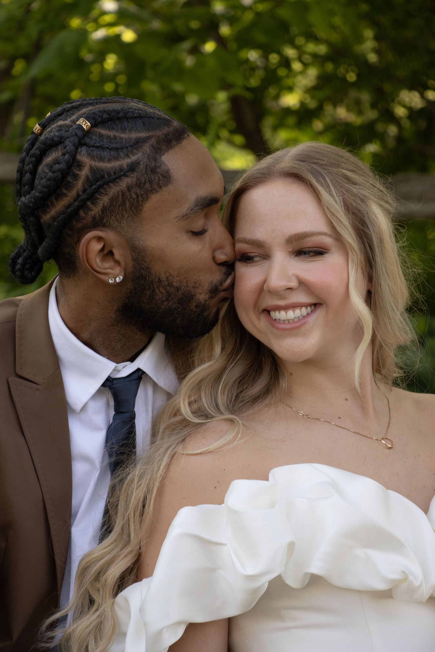 A man with braided hair in a brown suit and a woman in an off-shoulder white dress smile as the man kisses the woman on