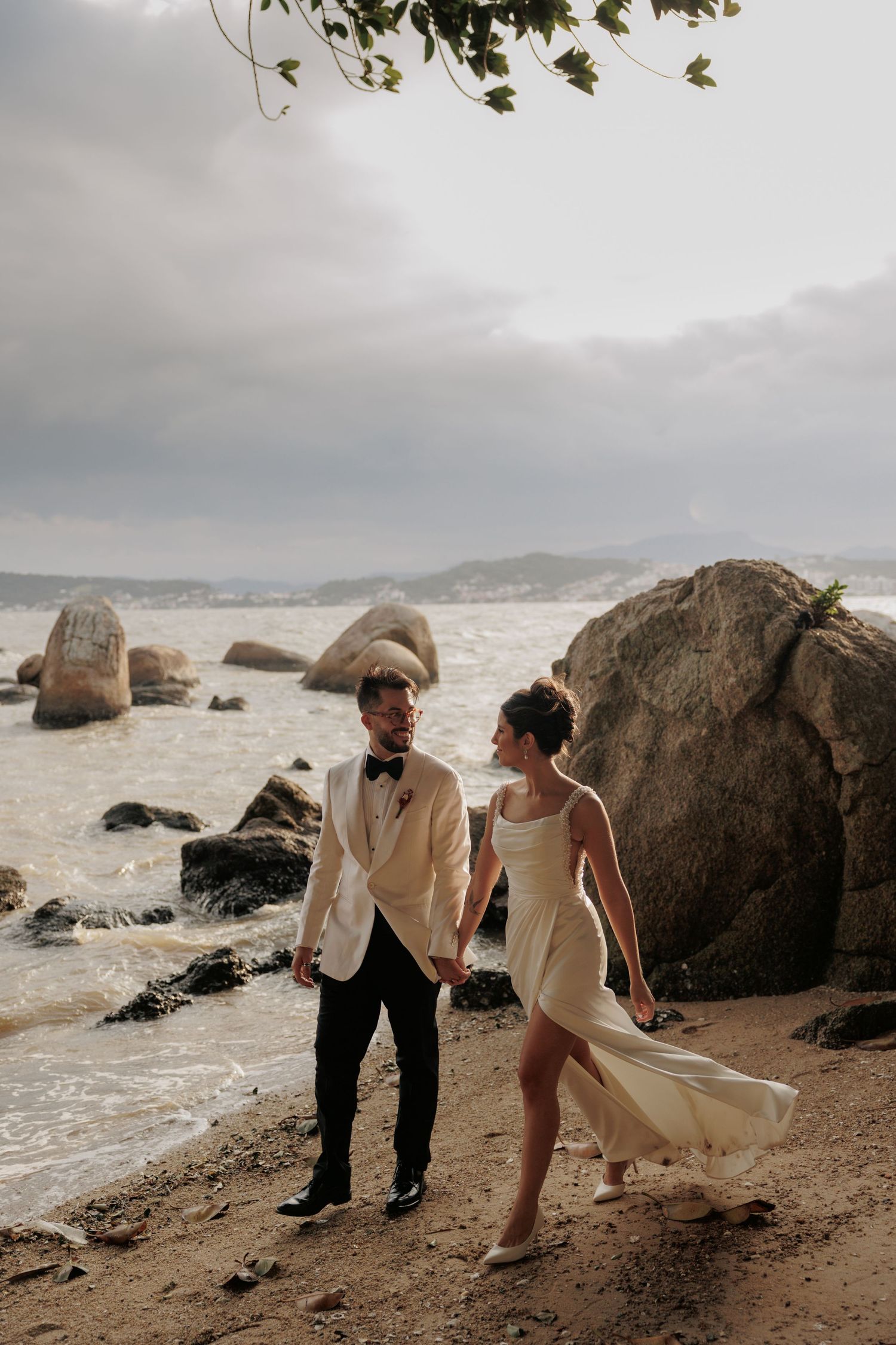 Couple in wedding attire walking on rocky coastal beach with ocean and mountains in background on overcast day.
