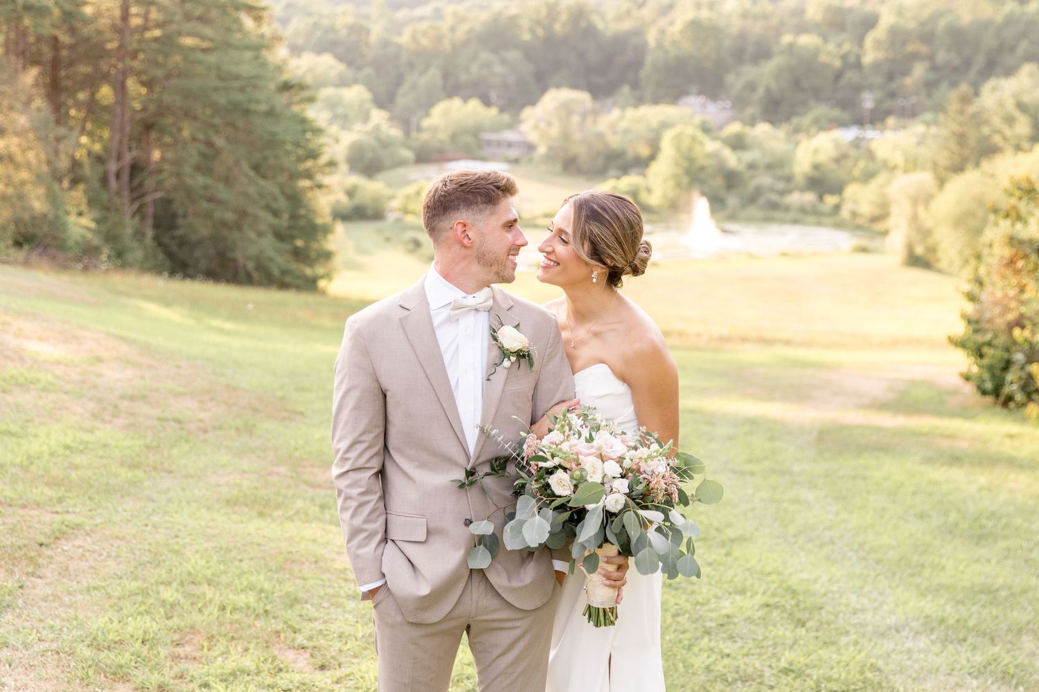 A couple in wedding attire embraces outdoors on a hillside with a beautiful floral bouquet in natural sunlight.