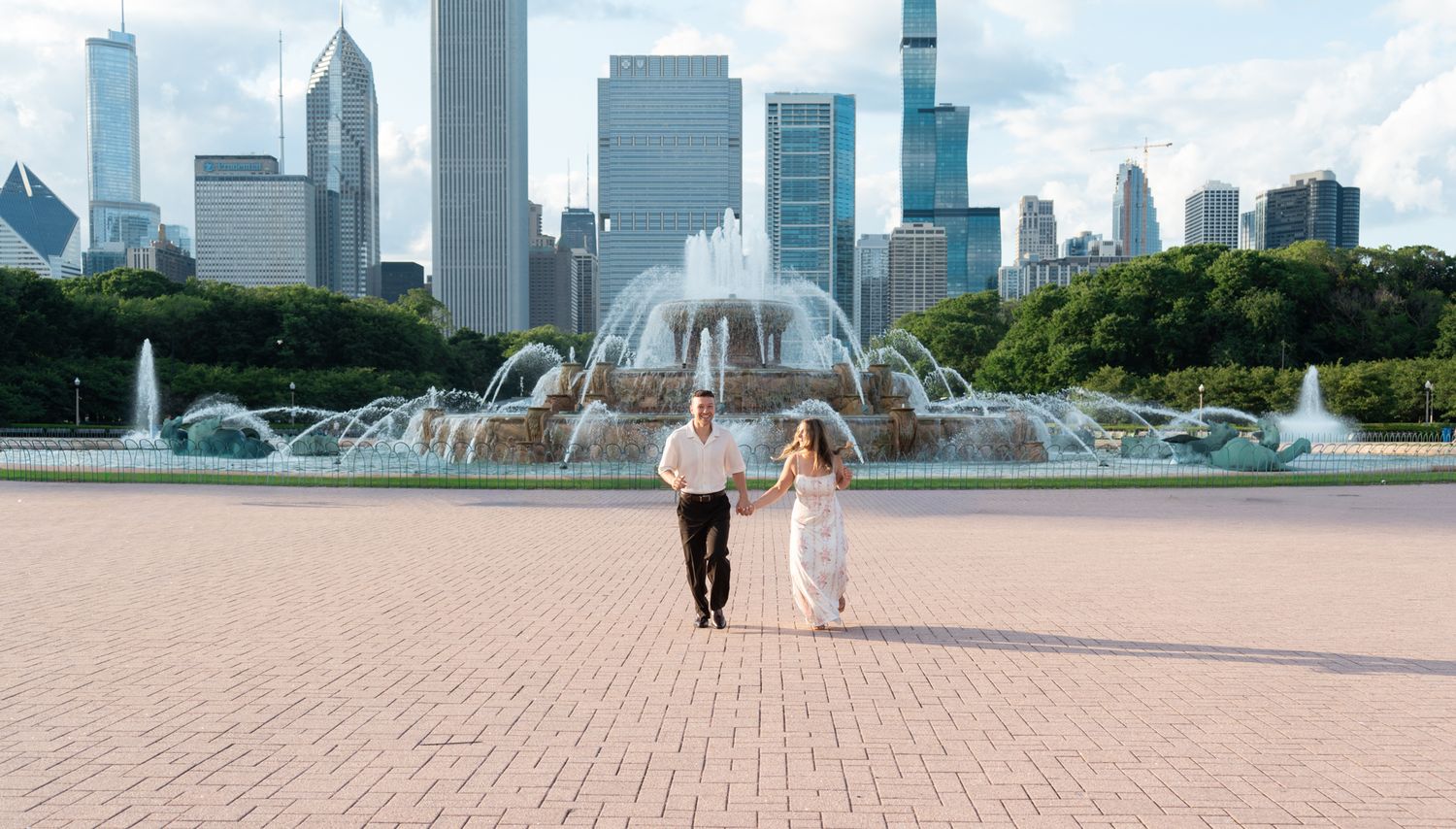 Couple in wedding attire holds hands running near Buckingham Fountain with Chicago skyline backdrop.