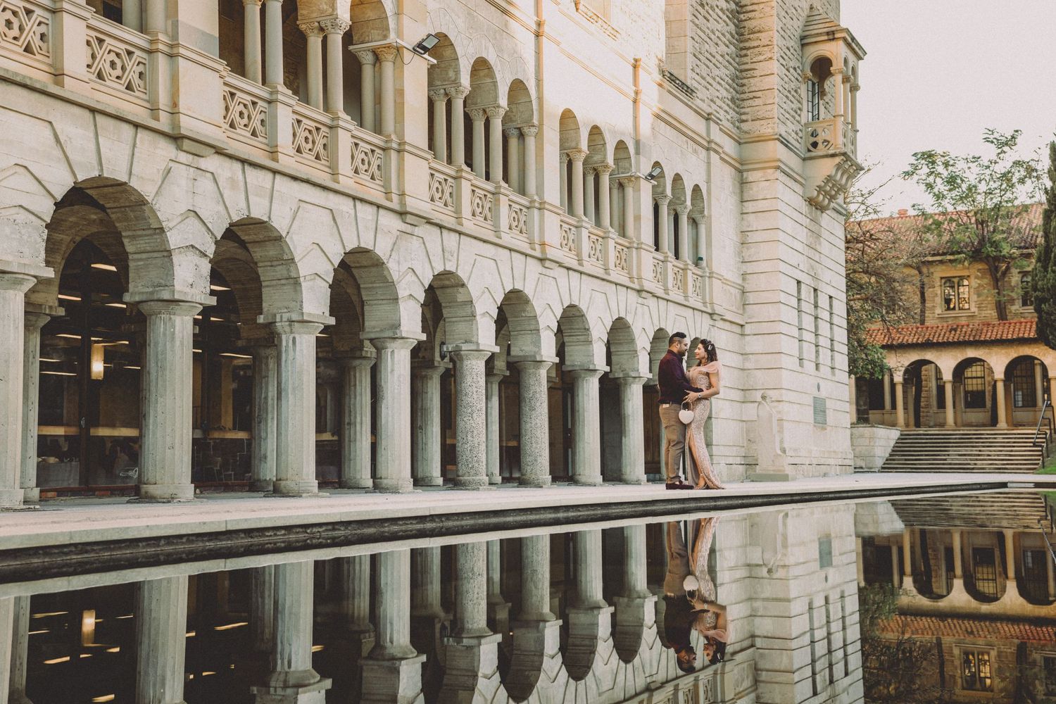 Couple sharing a romantic kiss by a reflective pool in front of a grand arched stone building.
