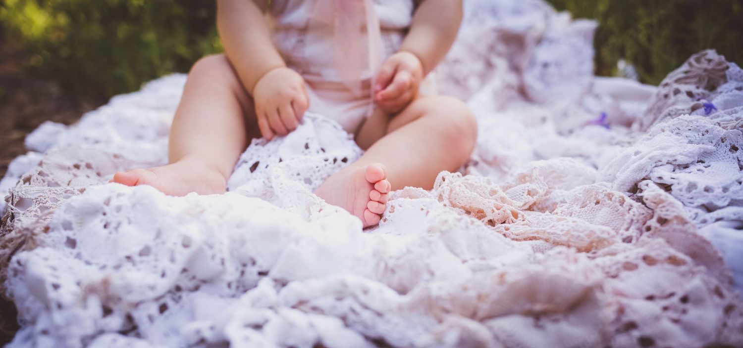 A baby sits on a lacy white blanket outdoors, wearing a pink outfit, with bare feet visible.