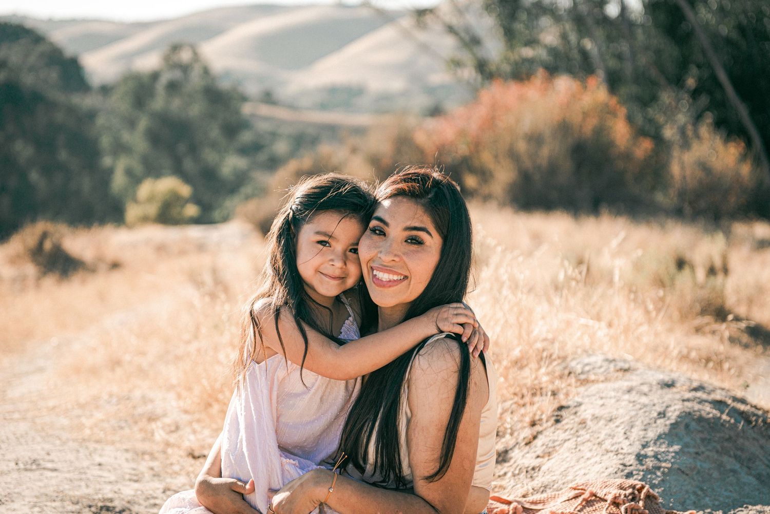 A mother and daughter smile warmly together on a sunny dirt path surrounded by golden hills and autumn foliage.