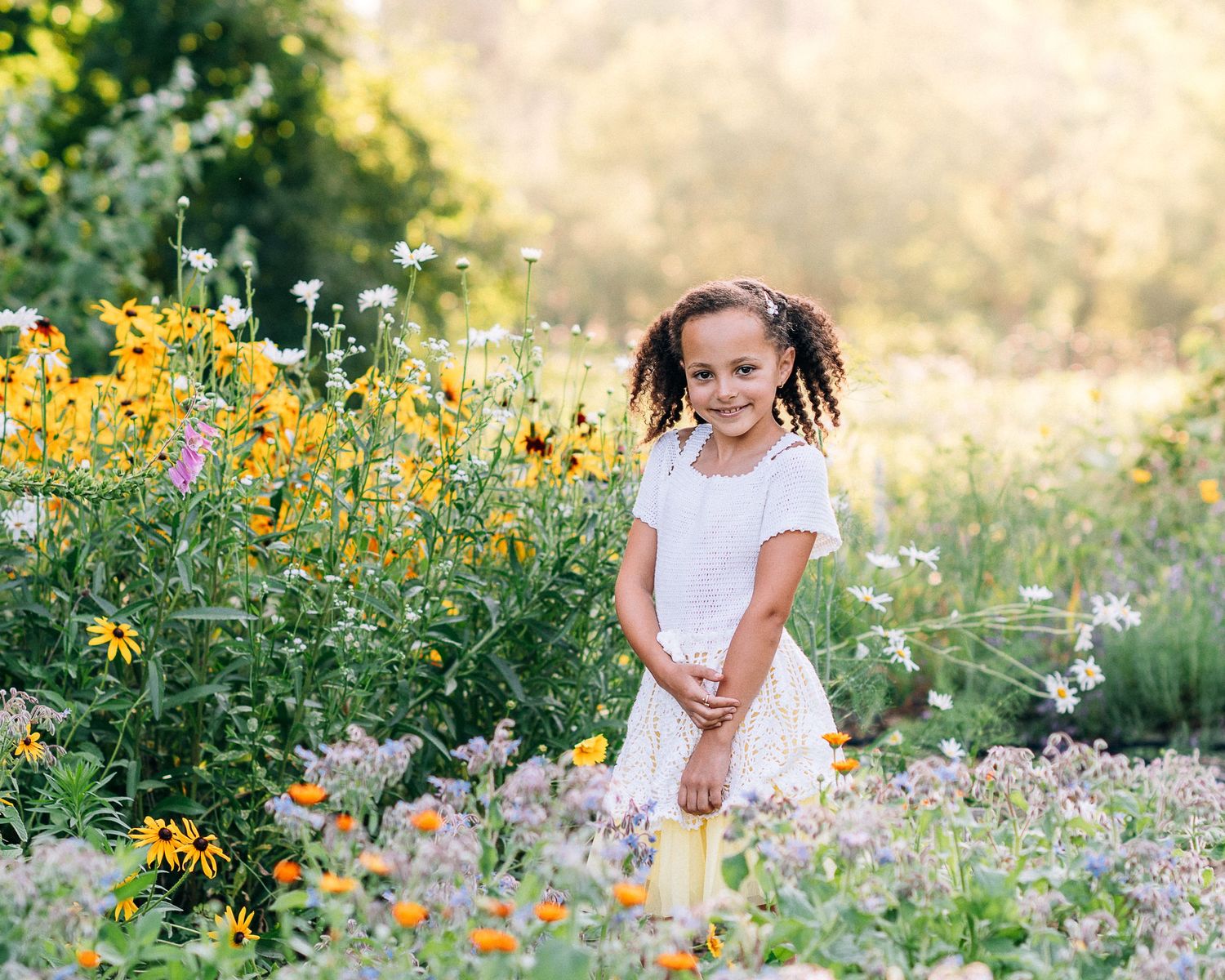 A young girl in a white dress smiles while standing among colorful wildflowers in a sunny garden.