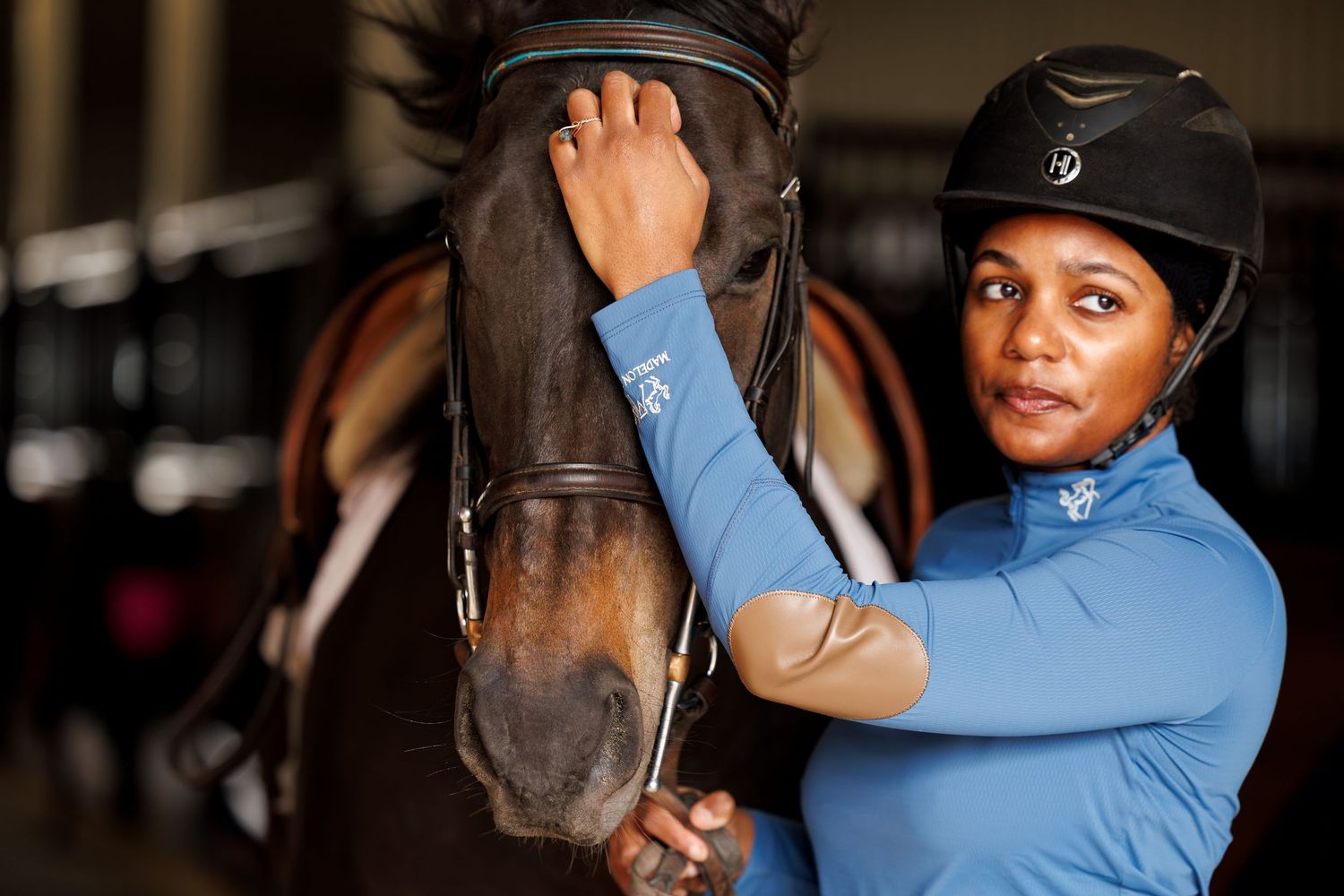 A young woman in a blue riding shirt and black helmet stands close to a dark bay horse in a stable.