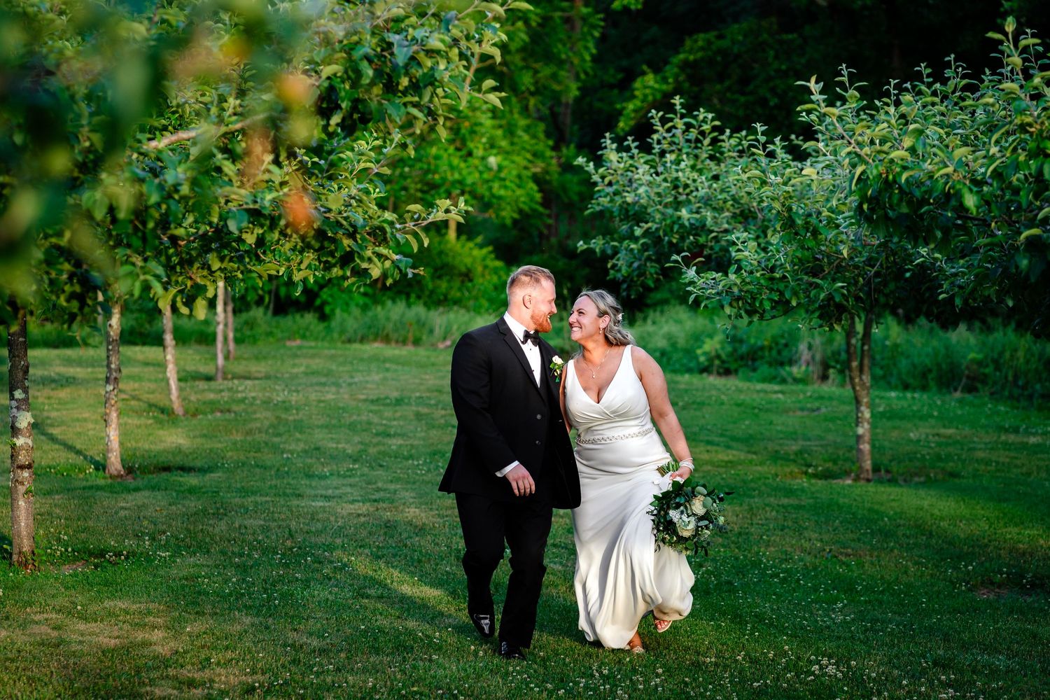 Bride and groom walking hand in hand through a lush green orchard on their wedding day.