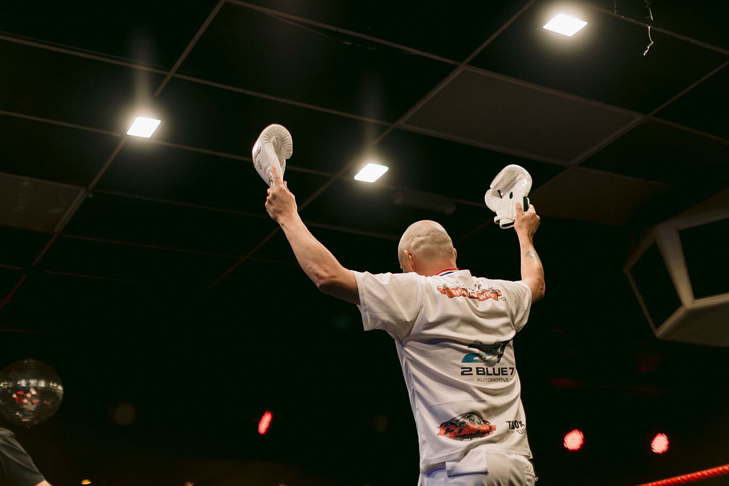 Bald boxer raises both arms in victory holding white towels in a boxing ring wearing a white t-shirt.