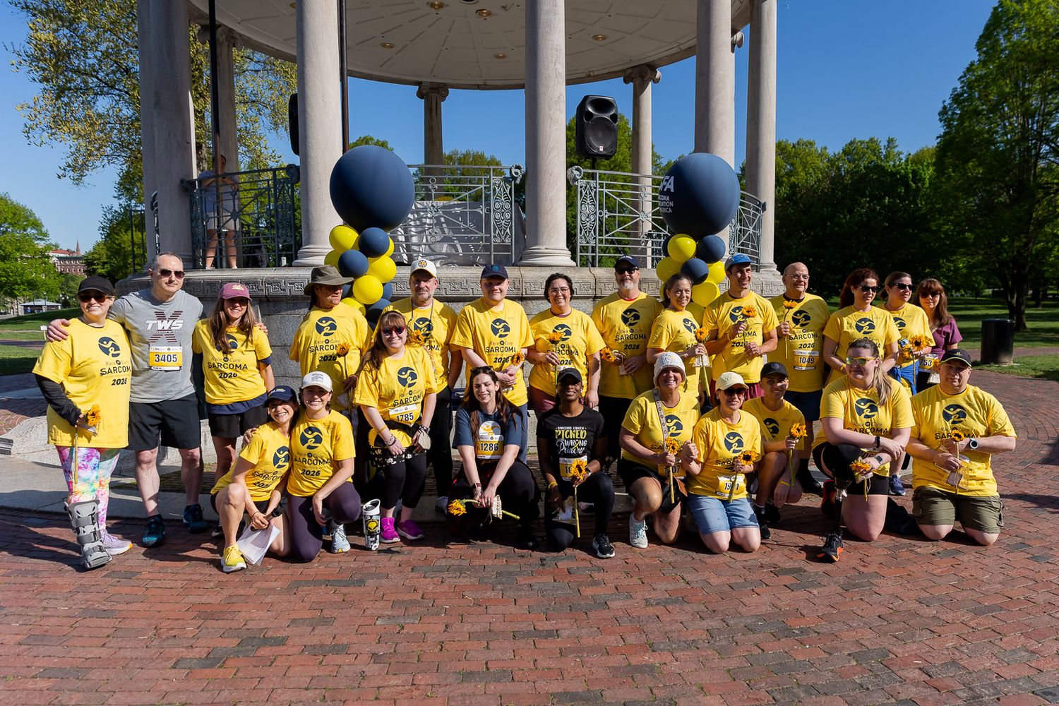 Large group in matching yellow shirts posing at a park gazebo with blue and yellow balloons on a sunny day.