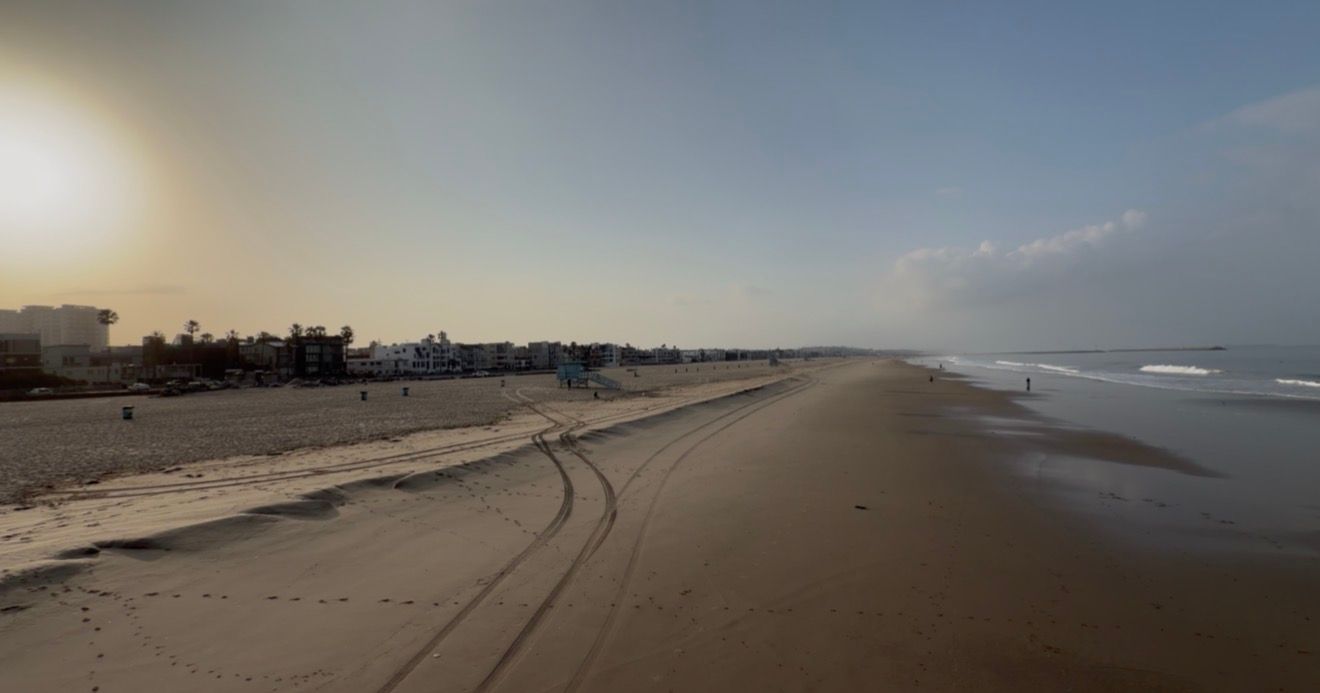Aerial view of a vast sandy beach at sunset with calm ocean waves and distant buildings along the shoreline.
