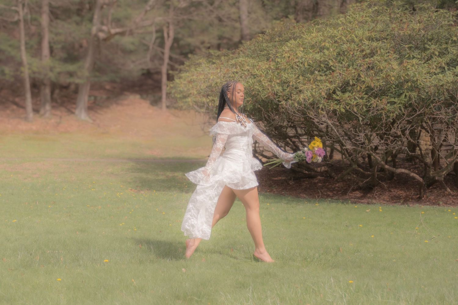 Woman in white lace dress running barefoot on green grass holding yellow flowers near bushes and trees.