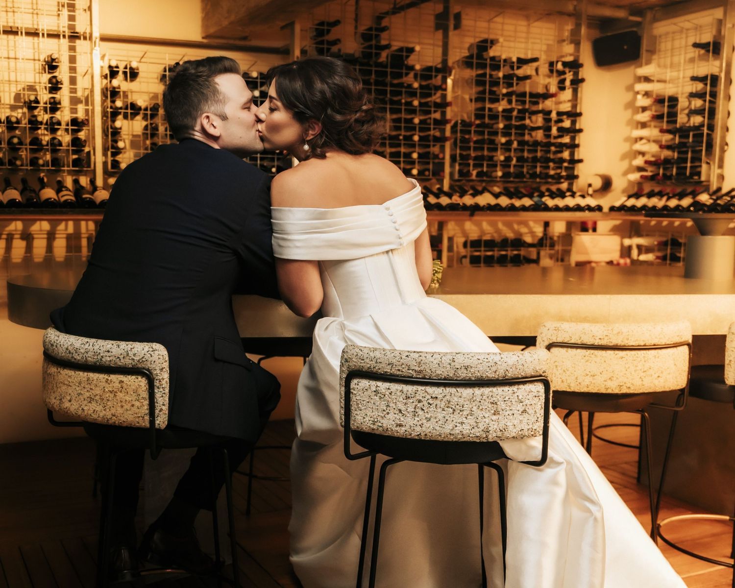 Bride and groom sharing a kiss at a romantic wine bar, dressed in elegant wedding attire.
