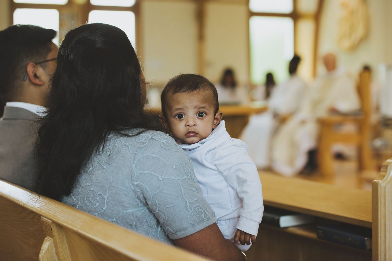 Baby in white outfit held by mother during a church baptism ceremony with priest visible in background.