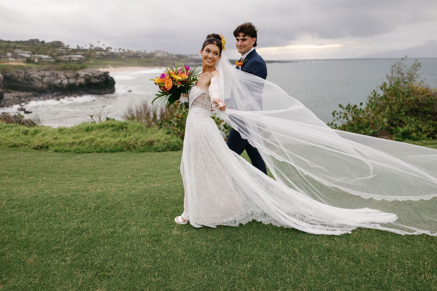 Bride and groom share an intimate moment on rocky coastal cliffs, bride holding vibrant tropical bouquet in lace gown and veil.