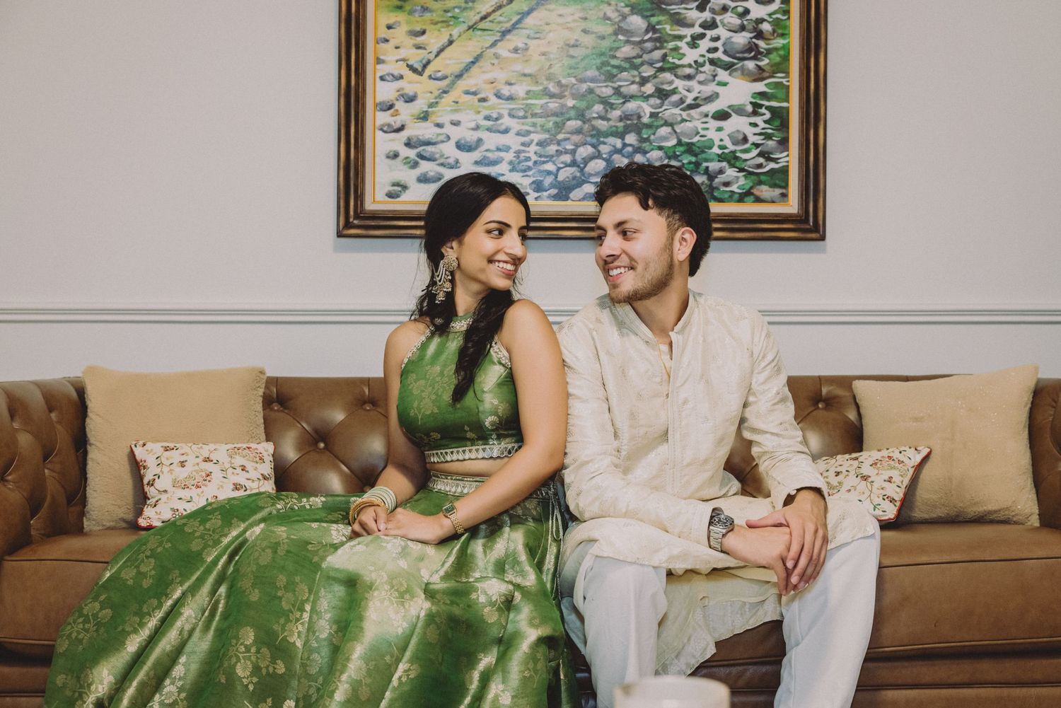 Couple in traditional South Asian attire sitting on a brown tufted sofa, smiling at each other.