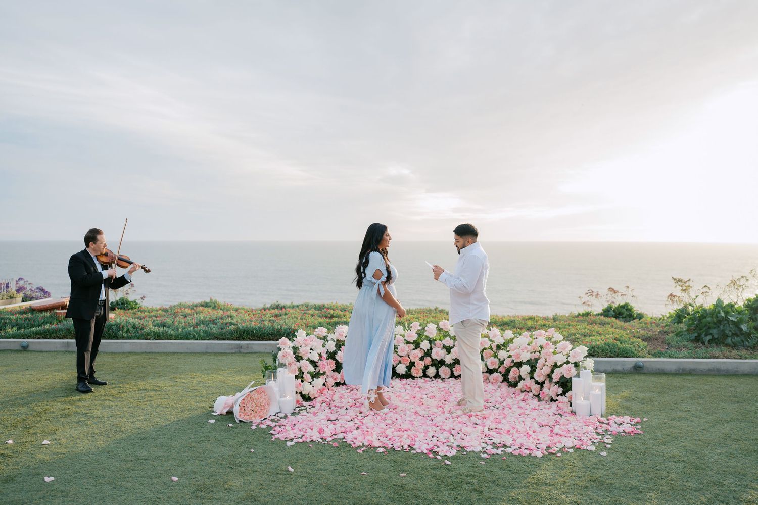 Romantic oceanside wedding ceremony on a cliff with pink rose petals and white floral arrangements at sunset.