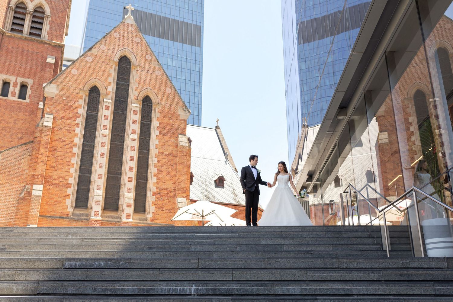 A bride and groom hold hands on sunlit steps between a historic brick church and modern glass buildings.
