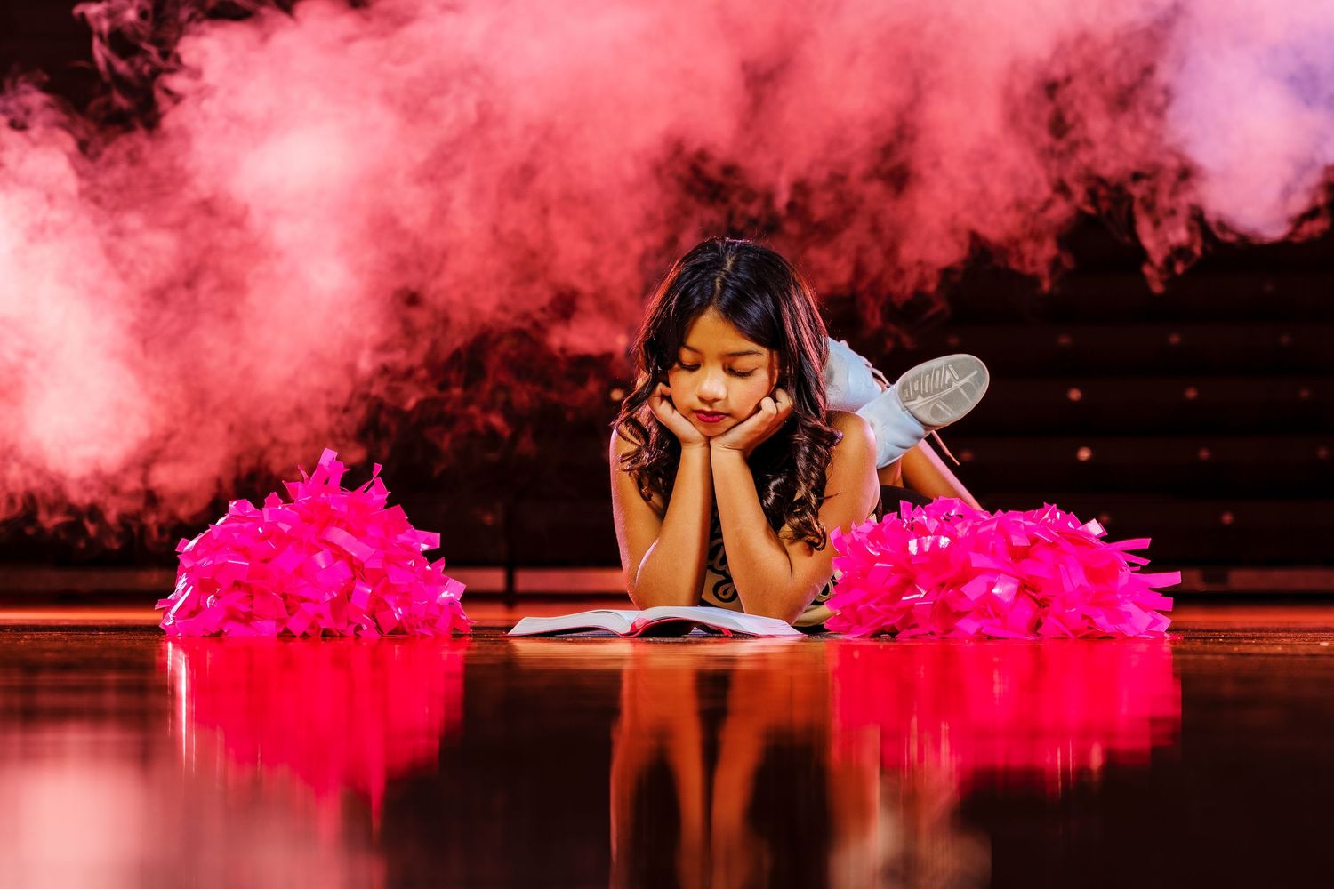 Young girl in fairy costume lying on stage floor surrounded by pink smoke and pompoms during a performance.