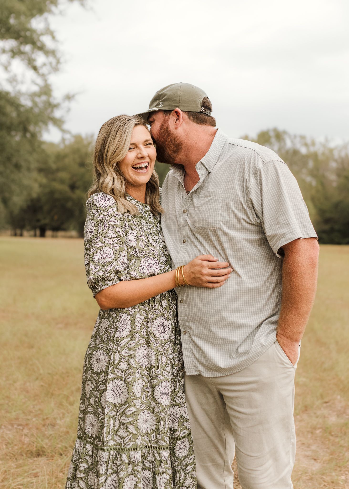 A laughing couple embraces in a green field, woman in floral dress, man in gray shirt and cap kissing her cheek.