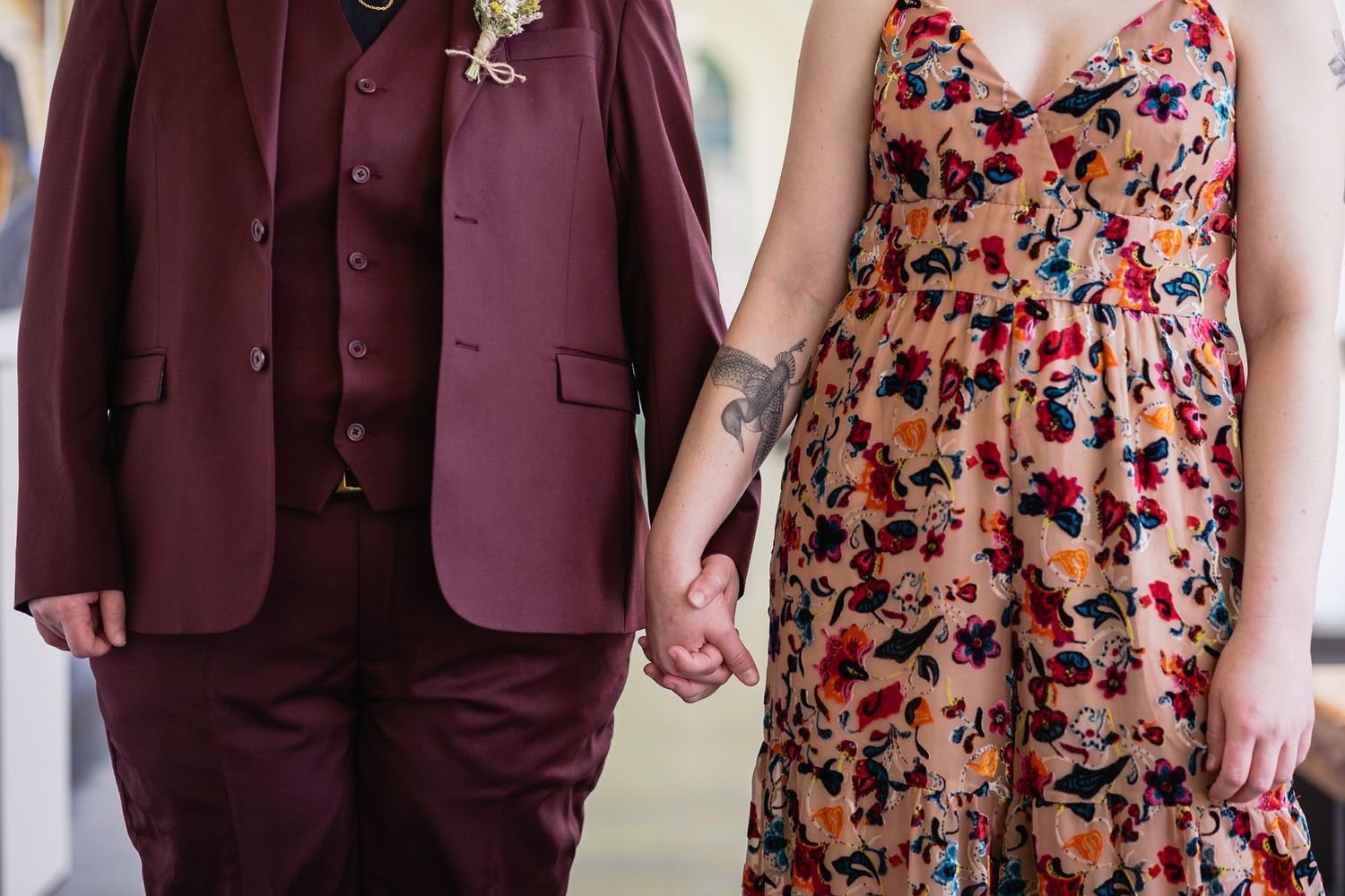Couple holding hands, one wearing a burgundy suit and the other a floral dress with a visible tattoo.