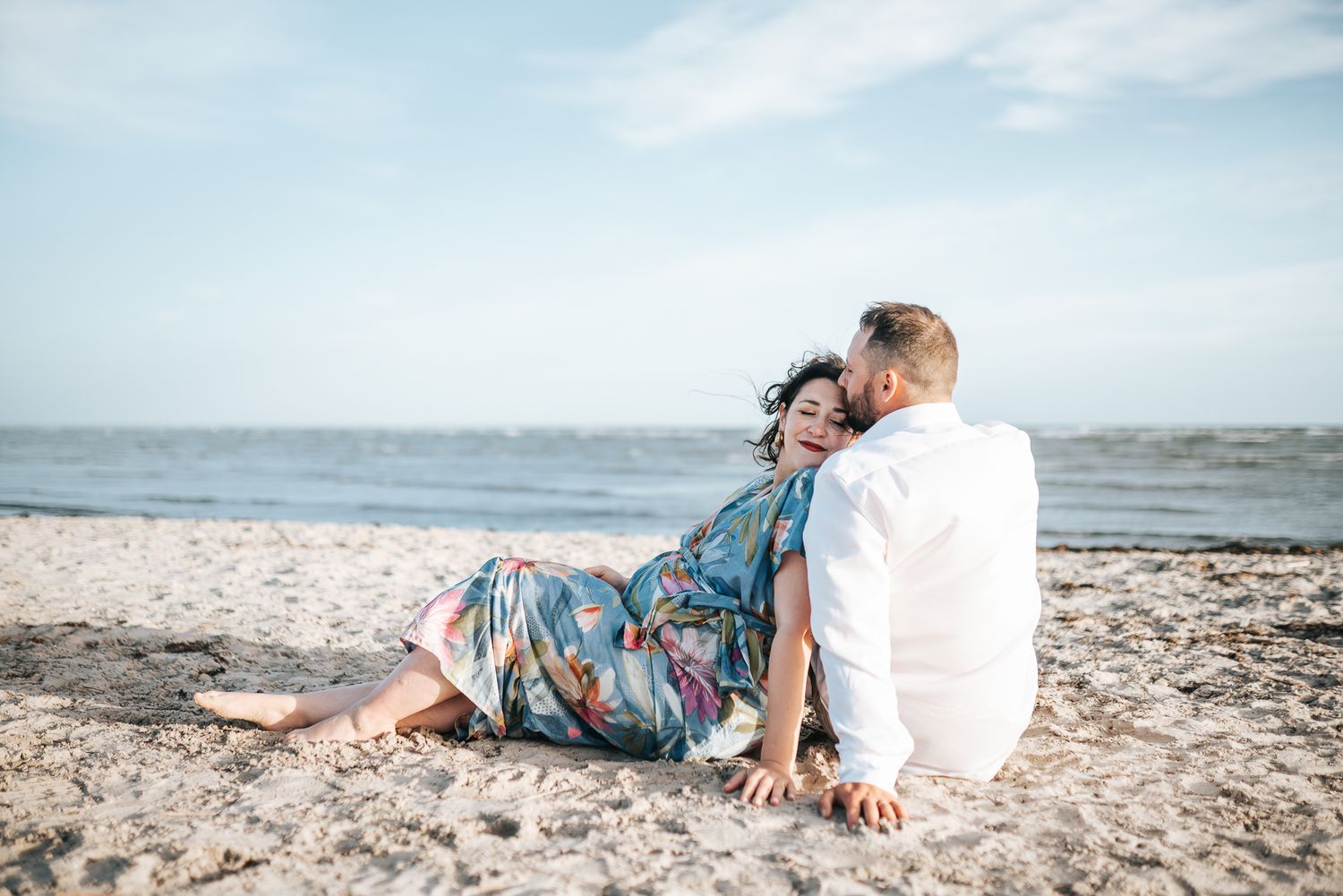 A pregnant couple in casual attire embracing on a sandy beach during a Melbourne sunset maternity photo session.