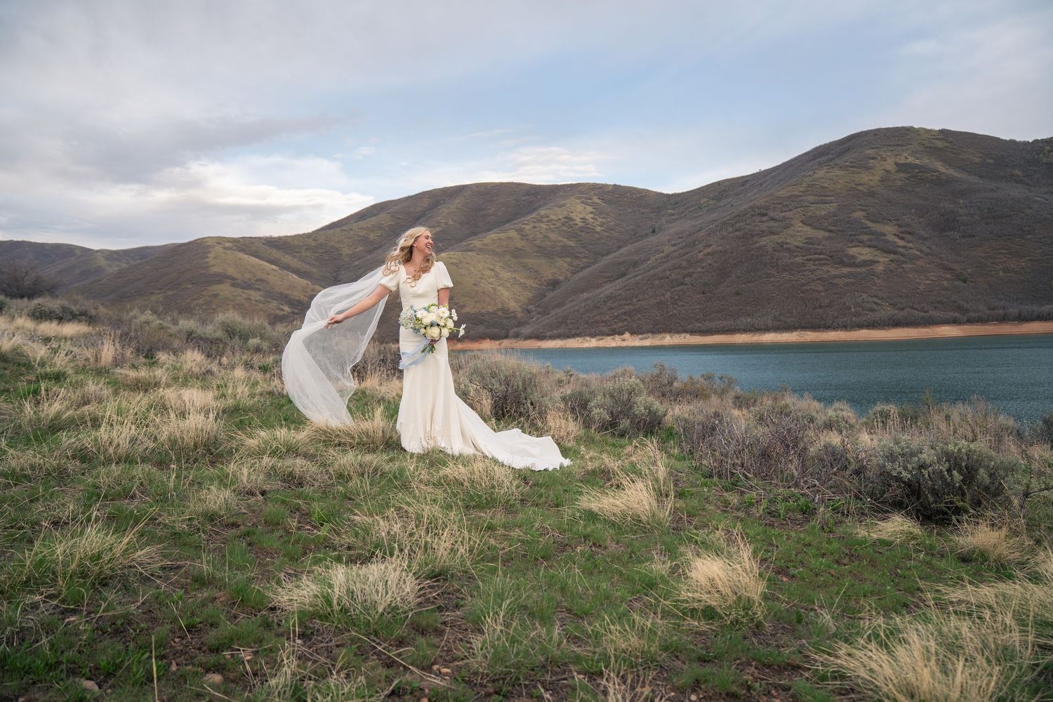 Bride in white gown with flowing veil stands on hillside near blue lake with mountain backdrop.